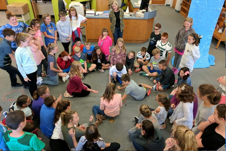 Students gather around for a championship-level match of marbles at the media center. Sixth grade students read a book about marbles and then played marbles to draw a connection.